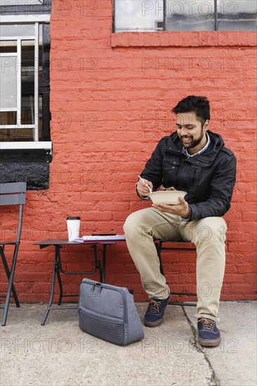 Hispanic man eating bowl of food on sidewalk