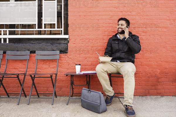 Hispanic man eating bowl of food on sidewalk using cell phone