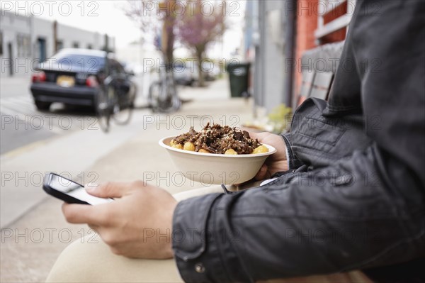 Hispanic man eating bowl of food on sidewalk using cell phone