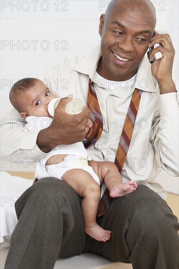 African father feeding baby daughter and talking on cell phone