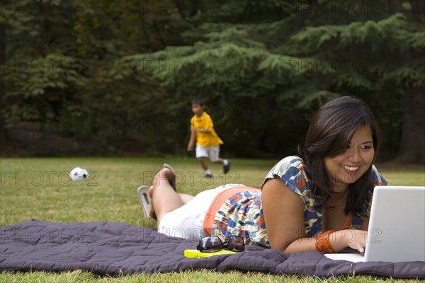 Asian mother using laptop in park with son in background