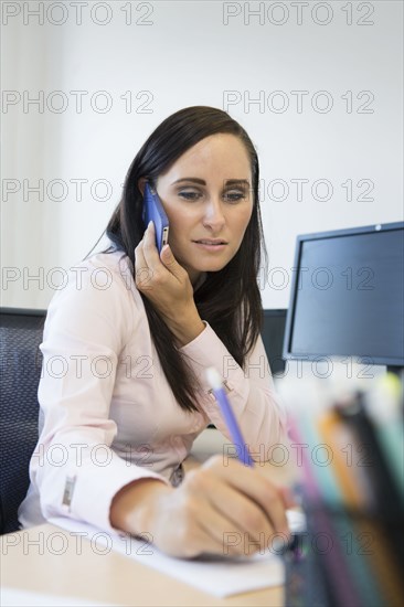 Caucasian businesswoman on cell phone taking notes at desk