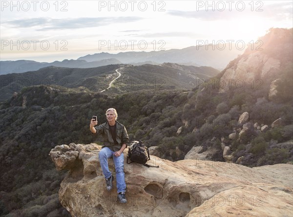 Older Caucasian man taking cell phone photograph on rocky hilltop