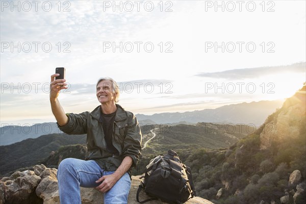 Older Caucasian man taking cell phone photograph on rocky hilltop