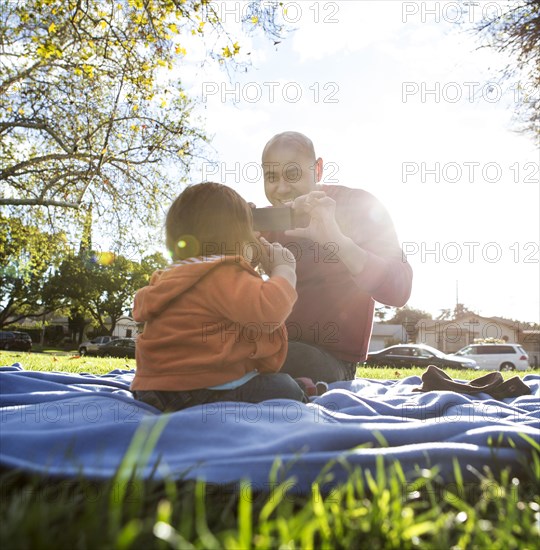 Hispanic father photographing son on blanket in park