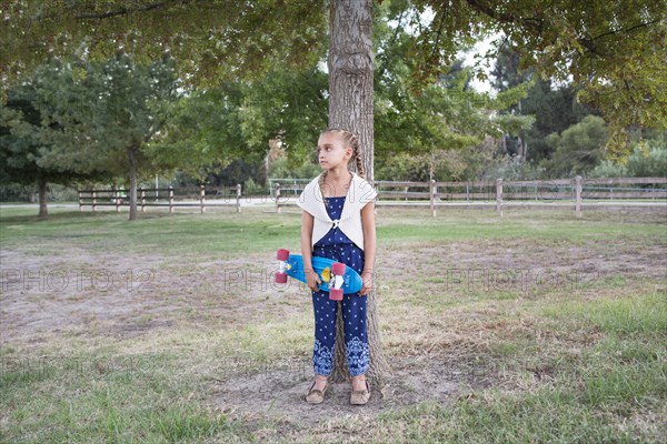 Mixed race girl holding skateboard at tree