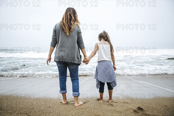 Hispanic mother and daughter standing barefoot on beach