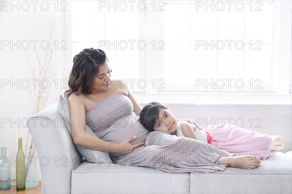 Hispanic girl listening to belly of pregnant mother on sofa