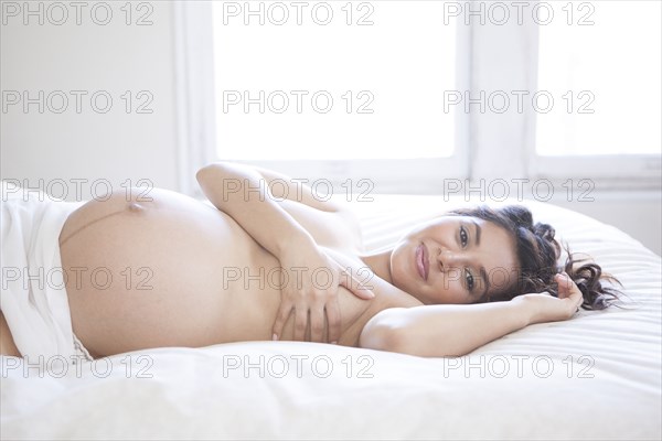 Nude Hispanic pregnant woman covering her breasts on bed