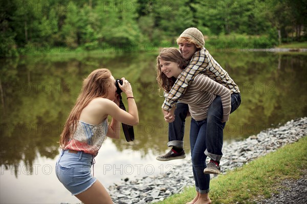 Girl photographing friend carrying boyfriend piggyback near lake