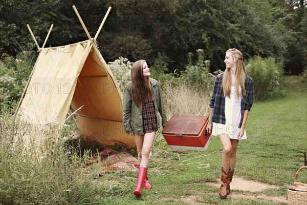 Women carrying vintage cooler near camping tent in forest