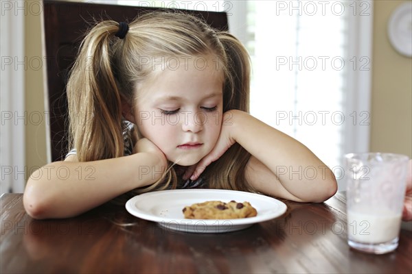 Girl staring at cookie with milk at table