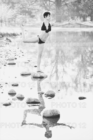Woman in lingerie practicing yoga on rock in still pond