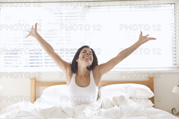Hispanic woman sitting in bed with arms raised