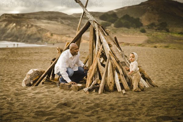 Father and baby son sitting under driftwood teepee on beach