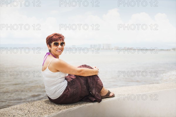 Hispanic woman sitting on stone wall overlooking beach