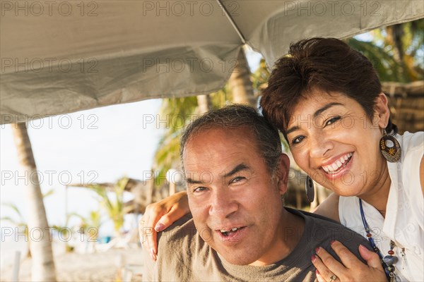 Hispanic couple smiling under umbrella on beach