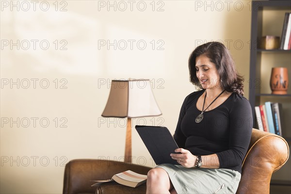 Hispanic woman sitting on armchair reading digital tablet