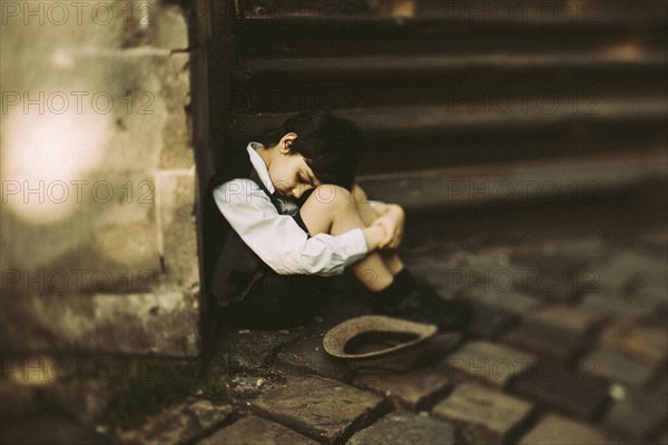 Old-fashioned Caucasian boy sitting near staircase begging for money