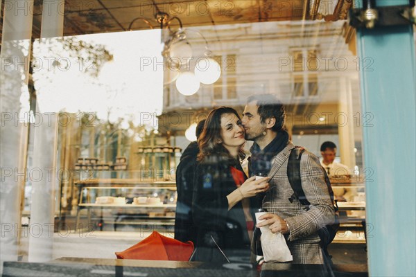 Caucasian man kissing woman on cheek behind bakery window