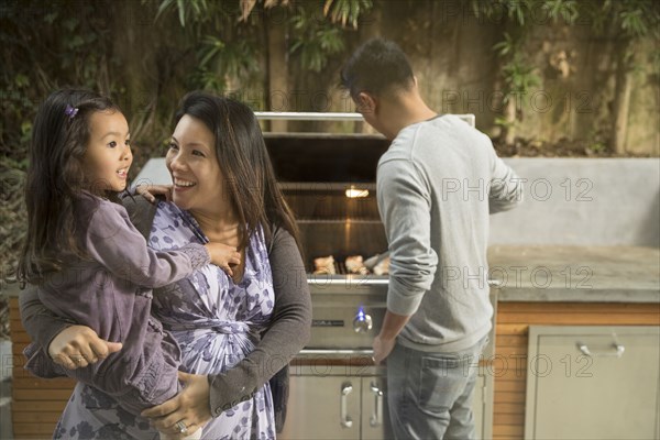 Expectant mother carrying daughter while husband is cooking on grille