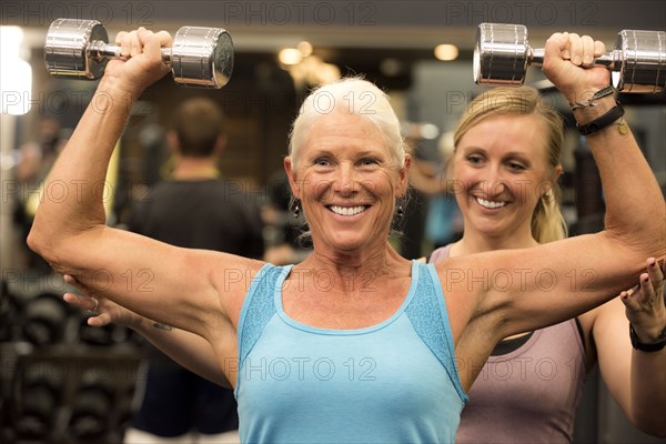 Trainer assisting older woman lifting dumbbells in gymnasium