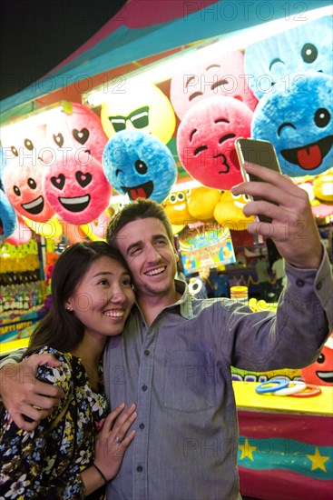 Smiling couple posing for cell phone selfie in amusement park