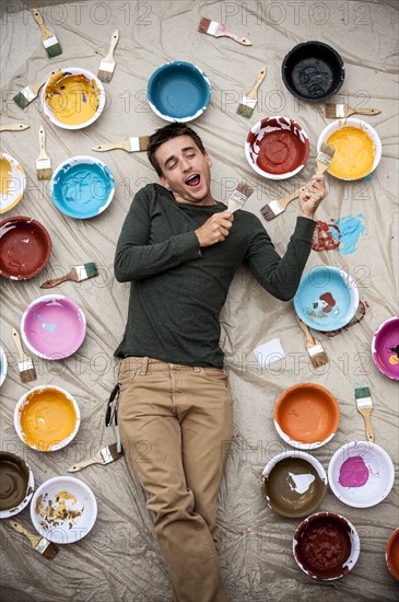 Man laying on tarp holding paintbrushes and singing