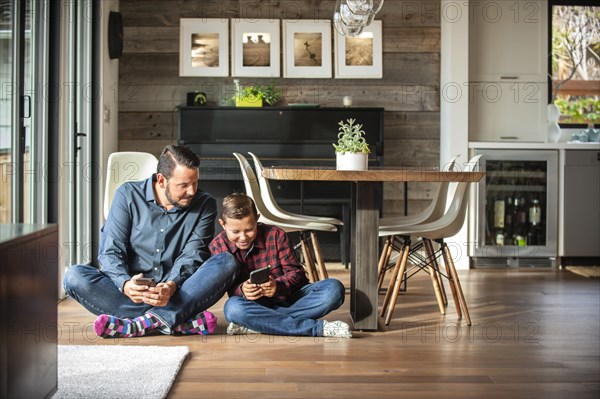 Smiling father and son sitting on floor texting on cell phones