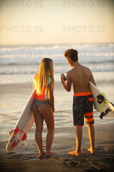 Teenage boy and girl holding surfboards texting on cell phone