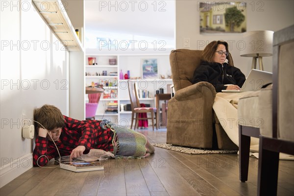 Caucasian boy laying on floor near mother using laptop in armchair