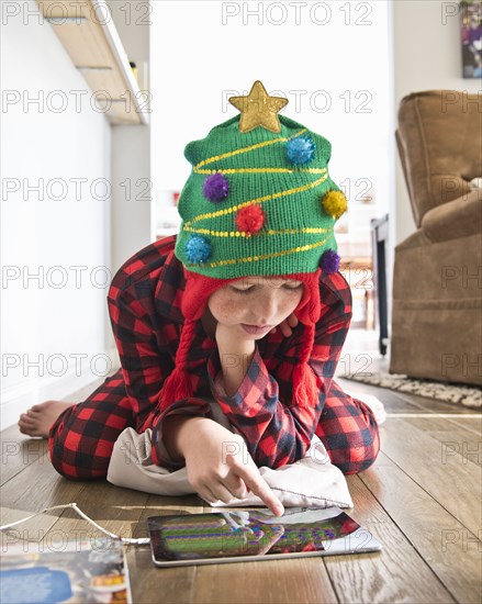 Caucasian boy wearing Christmas tree stocking-cap using digital tablet on floor