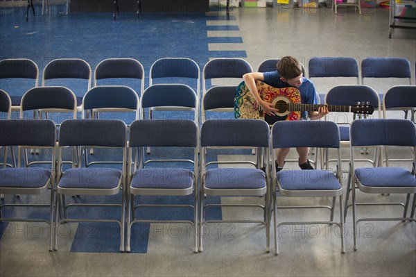 Caucasian boy sitting in row of chairs practicing guitar