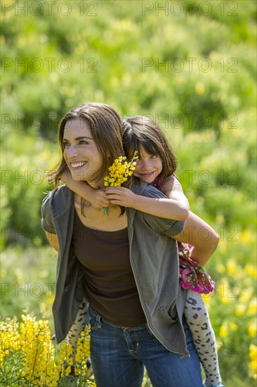 Caucasian mother carrying daughter piggyback on hillside with wildflowers