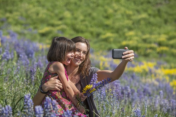 Caucasian mother and daughter posing for cell phone selfie on hillside with wildflowers