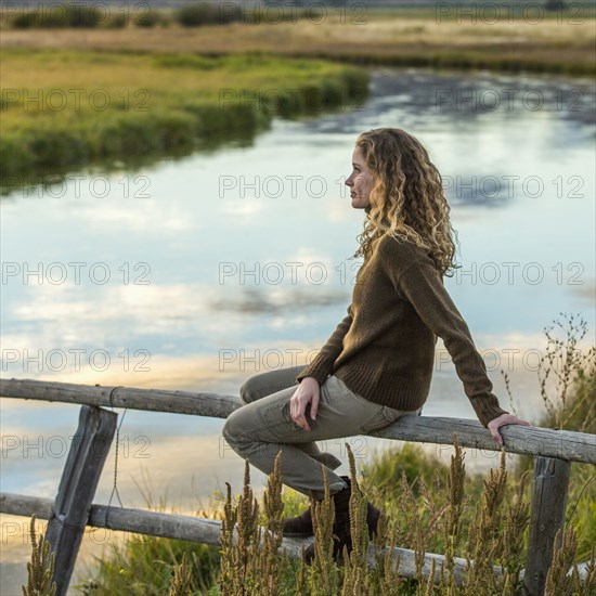 Caucasian woman sitting on wooden fence near river