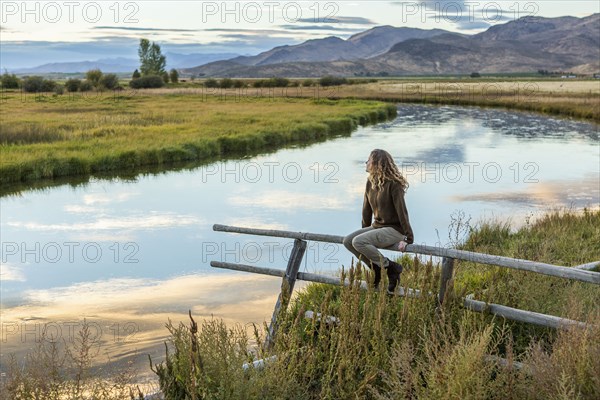 Caucasian woman sitting on wooden fence near mountain river