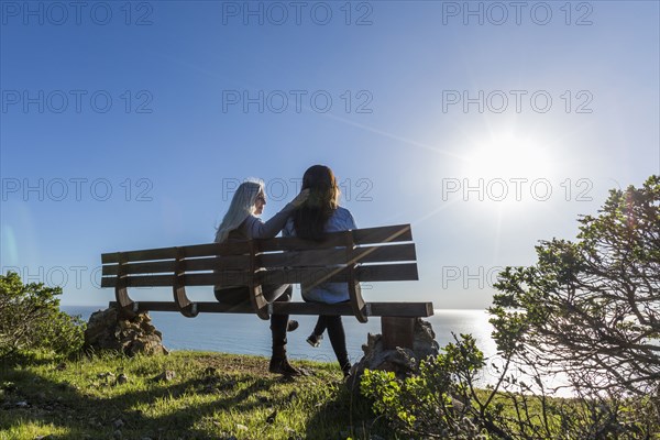 Caucasian mother and daughter sitting on bench near ocean