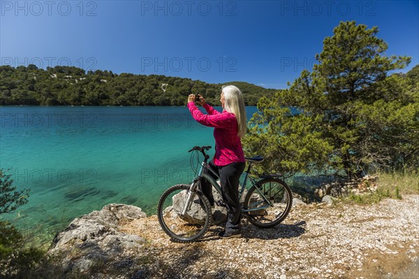 Caucasian woman on bicycle photographing lake with cell phone