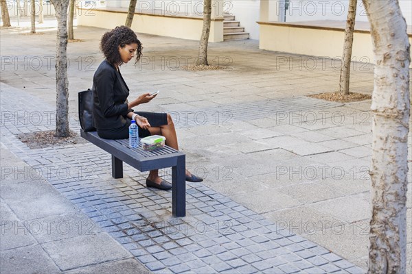 Mixed Race businesswoman eating lunch on bench texting on cell phone