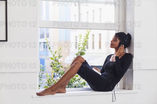 Mixed Race woman sitting in windowsill listening to headphones