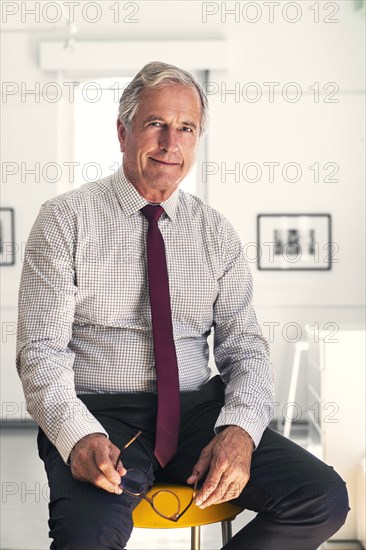 Smiling Caucasian businessman sitting on stool holding eyeglasses