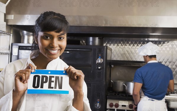 Mixed Race female chef holding Open sign in kitchen