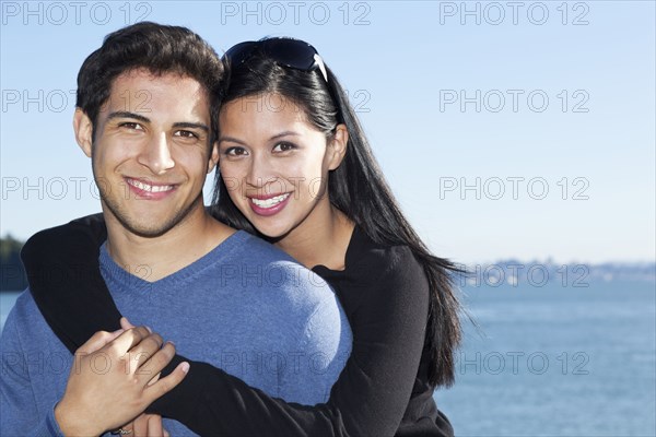 Smiling mixed race couple hugging by water