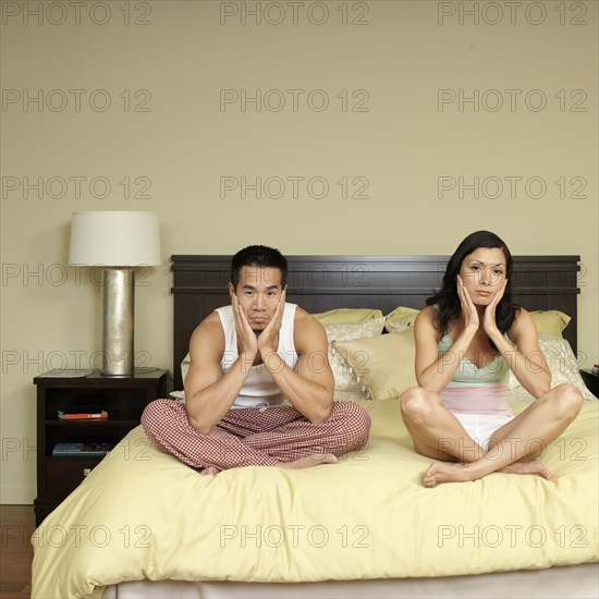 Sad Asian couple sitting cross-legged on bed