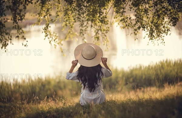 Caucasian woman sitting in grass near river holding hat