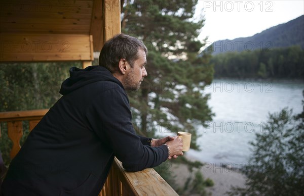 Caucasian man leaning on porch near lake drinking coffee