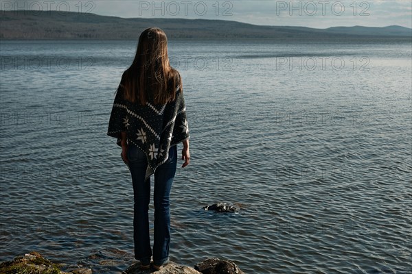 Pensive Caucasian girl standing on rock at lake