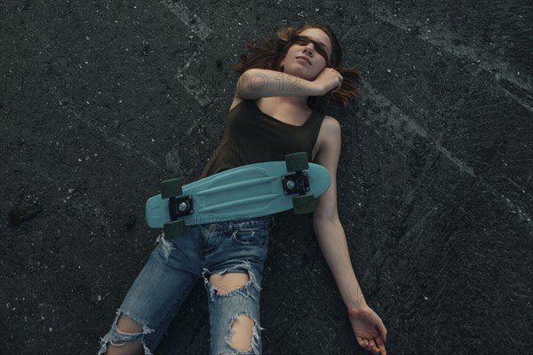 Caucasian teenage girl with skateboard laying on pavement covering eyes