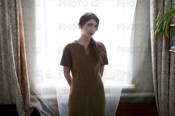 Serious Caucasian woman standing near window curtain